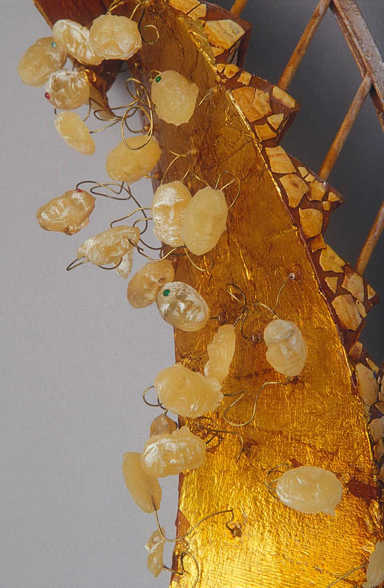 Translucent, amber-colored stones are suspended by coiled wires on a textured, gilt surface adorned with small, mosaic-like pieces. The background features a diagonal wooden lattice pattern. No text present.