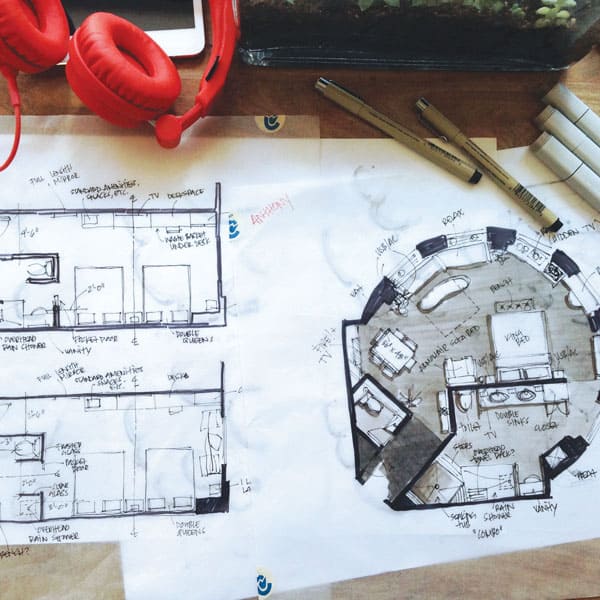 Architectural sketches lie on a wooden desk alongside red headphones, pens, and markers. The sketches detail room plans with labels like "double closet" and "laundry." A smartphone is partially visible.